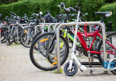 Zdjęcie przedstawia parking rowerowy ze stojakami typu odwrócone "U". Na parkingu licznie stoją rowery, a na pierwszym planie jest hulajnoga.