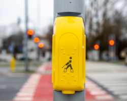 Zółty przycisk na szarym słupie sygnalizatora. Widok z przodu. Widoczny symbol pieszego na przejściu oraz napis w języku Braille'a.