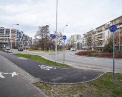 Na zdjęciu widać chodnik, drogę rowerową, ulice, bloki mieszkalne, dwie kobiety idące z wózkami i przejeżdżające samochody