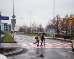 Uczniowie wracają ze szkoły, przemierzają pasy drogowe,, jeden z nich jedzie na hulajnodze