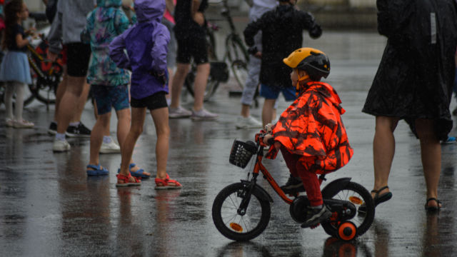 Dziecko jedzie rowerem w deszczowy dzień, w tle widać tłumnie zgromadzoną młodzież