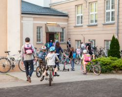 Dzieci zmierzające na zajęcia na rowerach i hulajnogach, w tle budynek szkoły