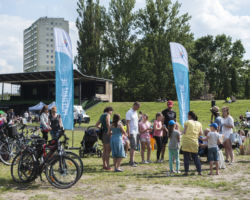 Uczestnicy pikniku na zakończenie kampanii Rowerowy Maj 2016 na Stadionie Syrenki, na zdjęciu widać rowery, flagi z napisem "Przesiądź się", dorosłych i dzieci, w tle drzewa i bloki