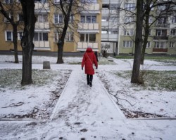 Kobieta idąca ośnieżonym chodnikiem, w tle bloki mieszkalne.